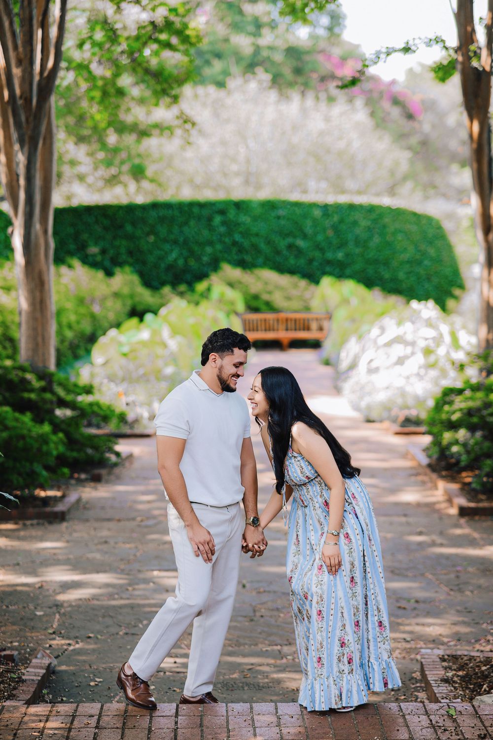 A couple holds hands and laughs together on a brick garden path surrounded by lush greenery and blooming trees.