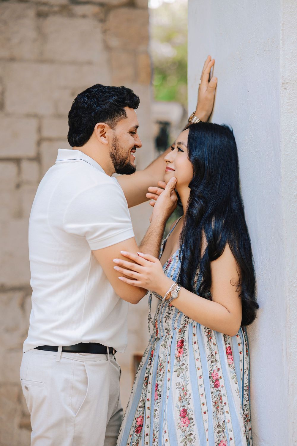 A romantic couple shares a tender moment, with a man in a white polo gently touching a woman's face against a stone wall.
