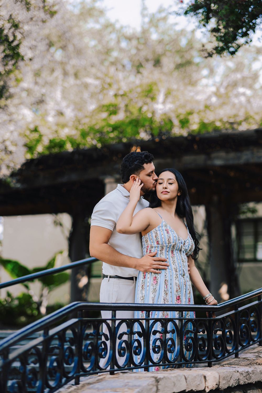 Romantic couple embracing on ornate iron staircase with blooming trees in background during outdoor engagement photo session.