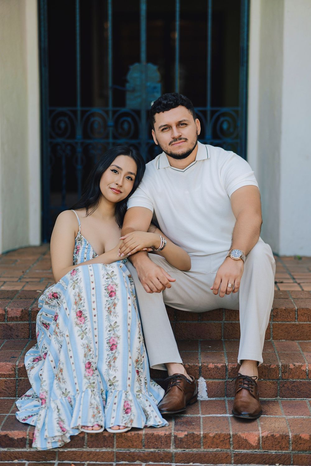 A couple sits on brick steps, woman in floral dress, man in white shirt and khakis, posing together outdoors.