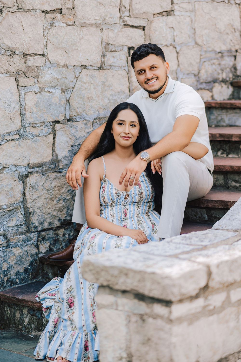 A couple poses on stone steps, woman in floral dress, man in white shirt, smiling warmly against a stone wall backdrop.