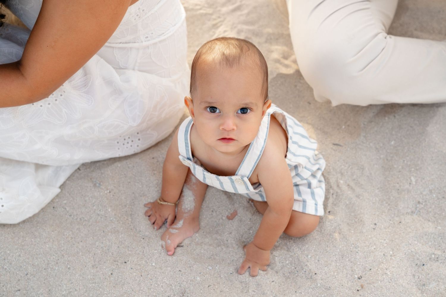 Curious baby in striped romper crawling on sandy beach, looking up at camera with bright eyes.