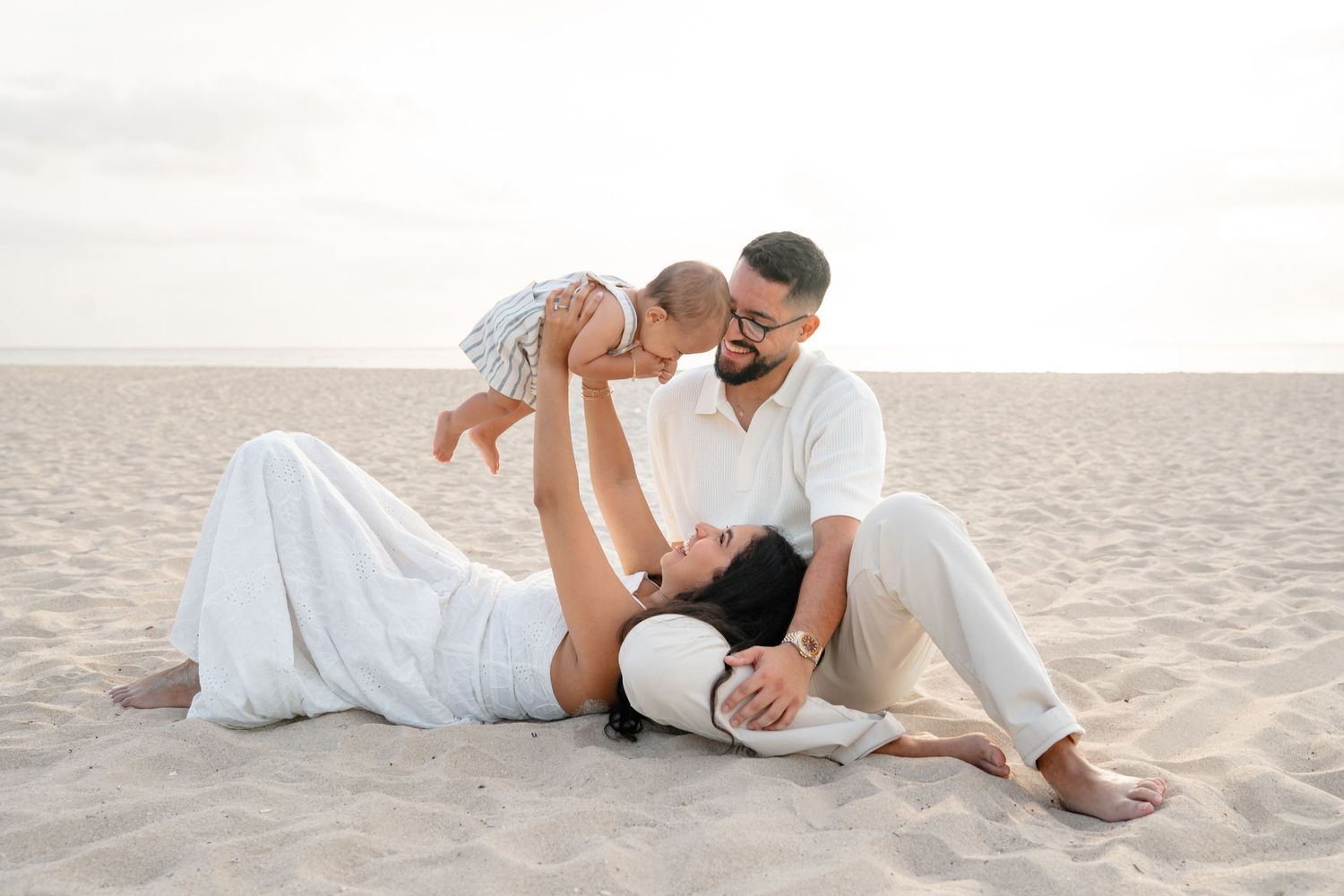 Family of three enjoying a beach day, parents sitting on sand lifting their baby in the air, dressed in white outfits.