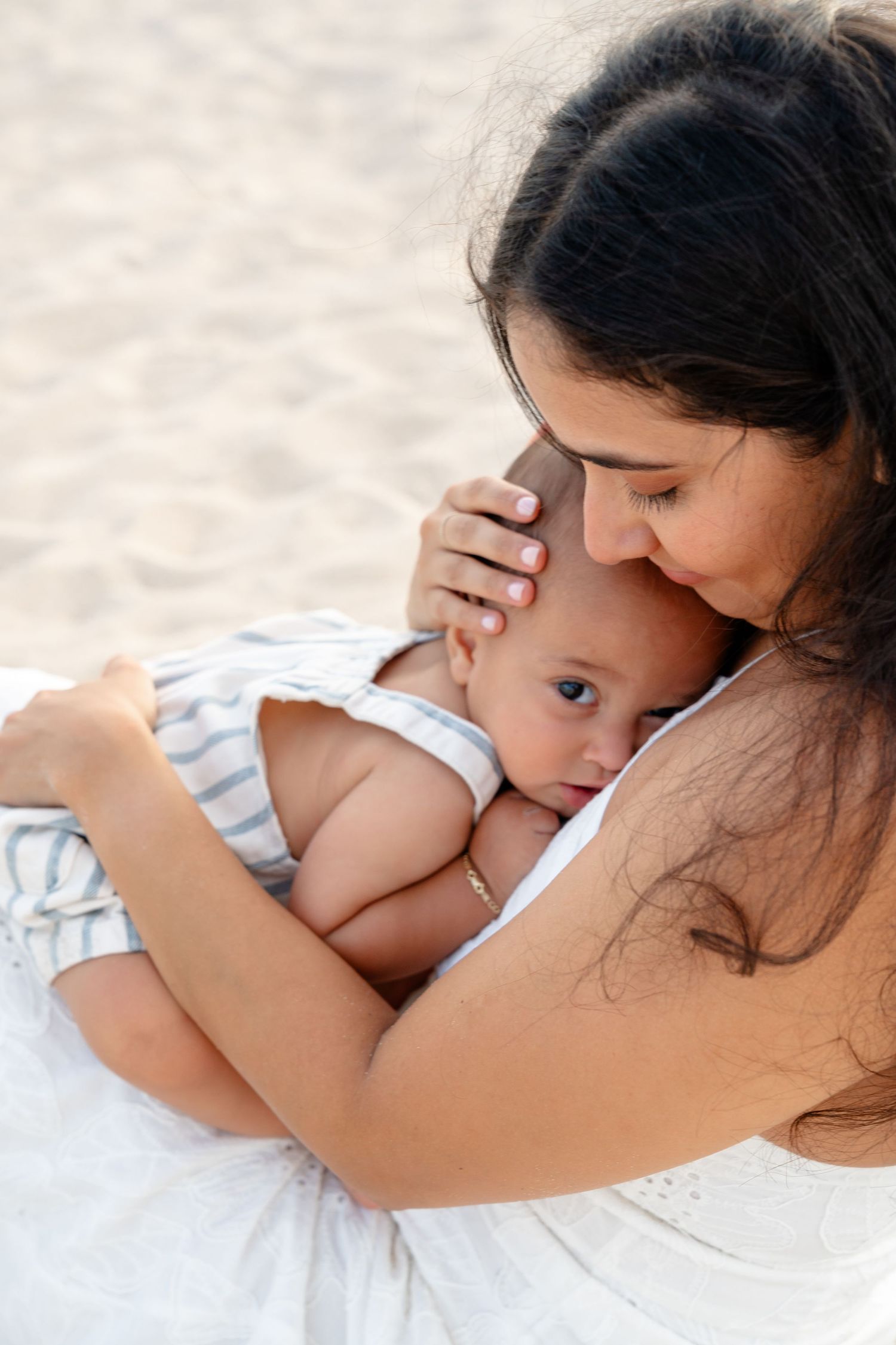 A mother tenderly kisses her baby's forehead while holding them close on a sandy beach.
