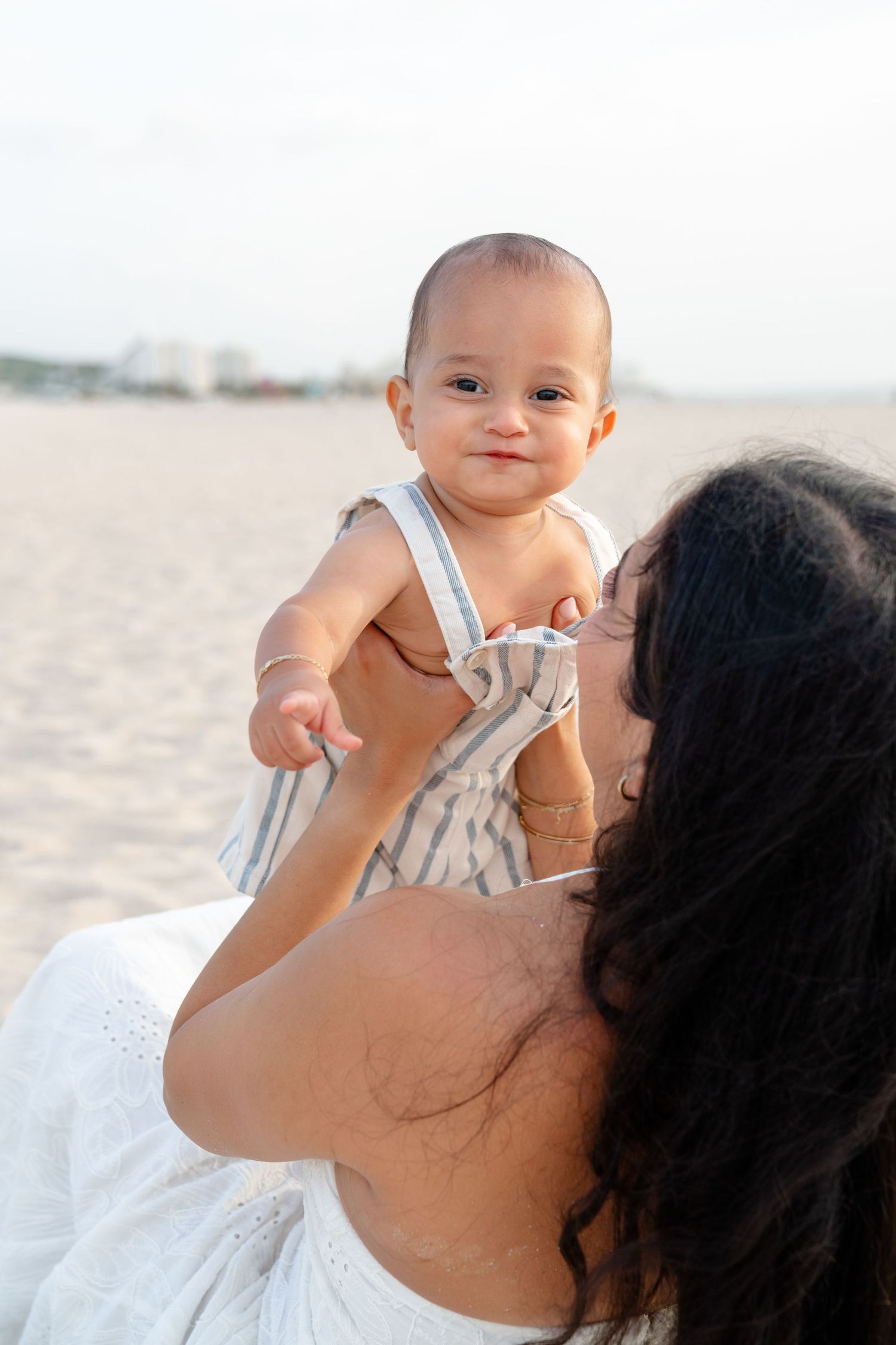 A mother lifts her smiling baby on a sandy beach, both dressed in white and stripes on a sunny day.