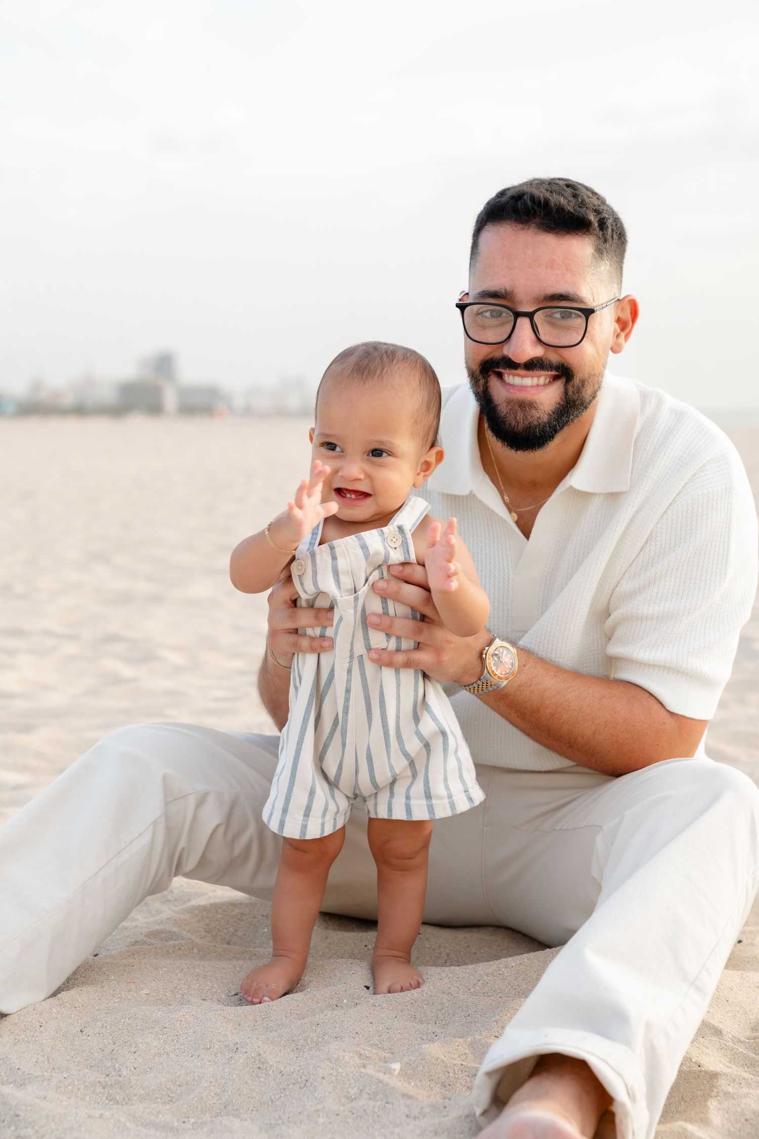 Smiling bearded father with glasses holds baby standing on sandy beach, both wearing light-colored outfits.