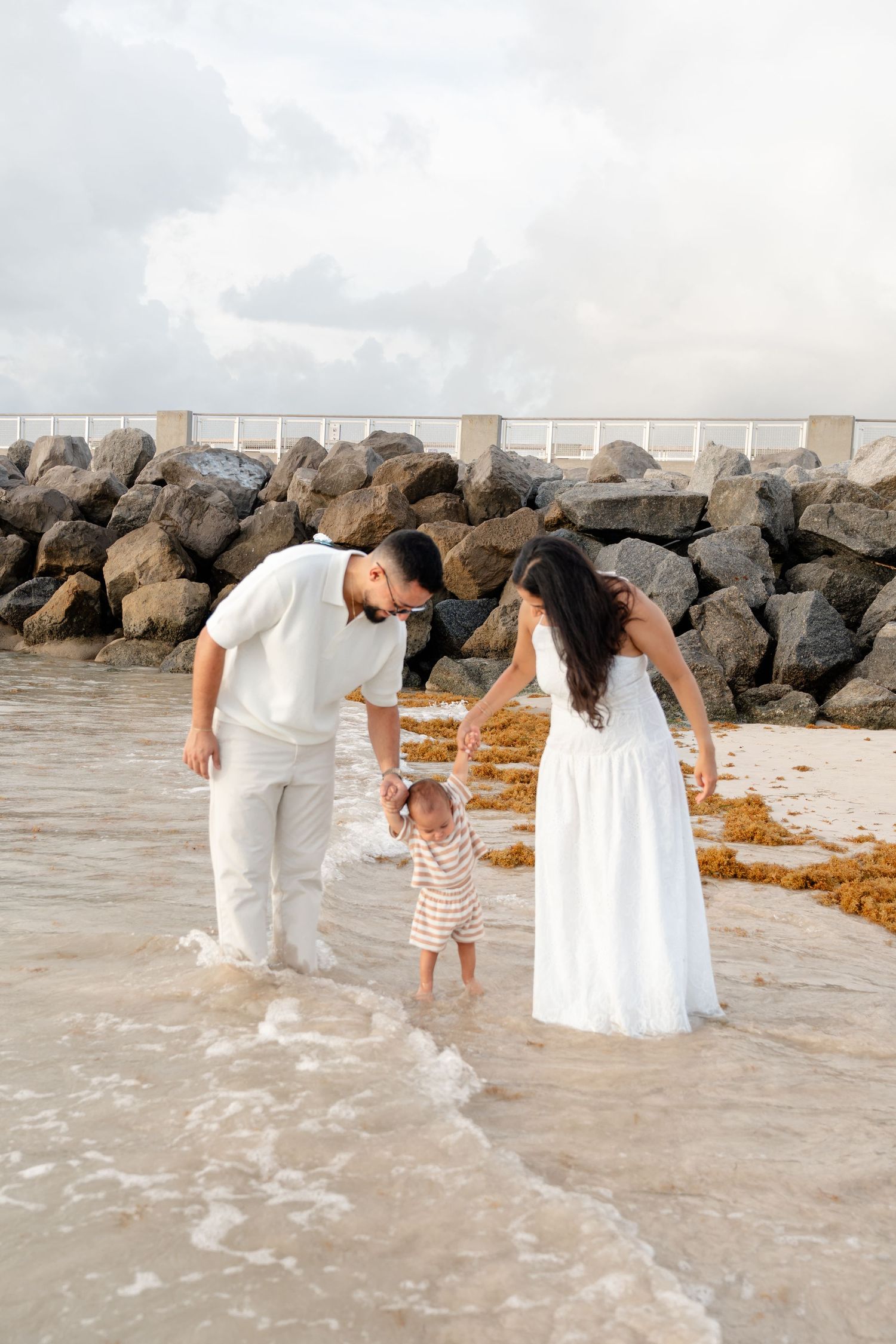 Family in white outfits playing in ocean waves on a rocky beach, parents holding toddler's hands.