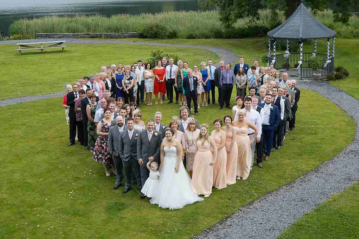 Wedding group photo arranged in heart shape on green lawn with bride, groom, and guests at outdoor venue.
