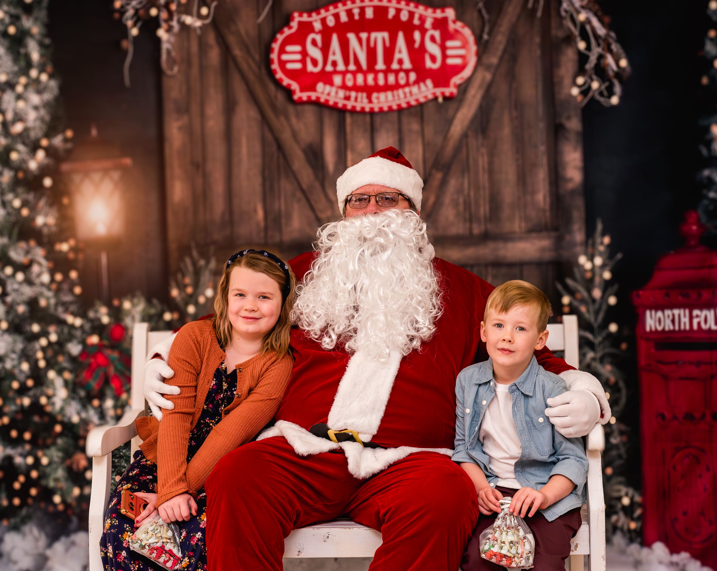 Two children sit with Santa Claus in his workshop, surrounded by festive Christmas decorations and trees.