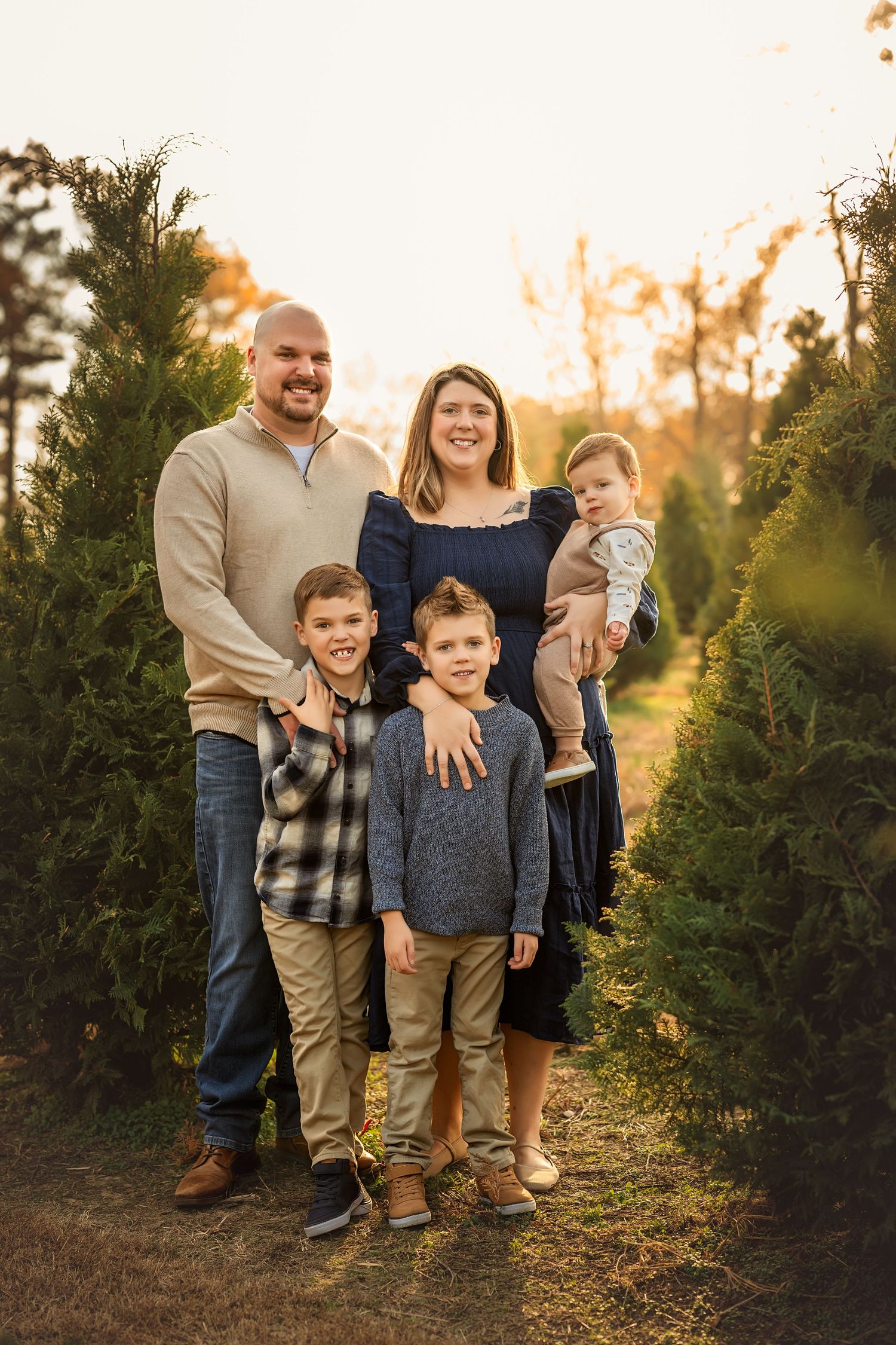 Family of five posing among Christmas trees at golden hour, parents with three young children smiling outdoors.