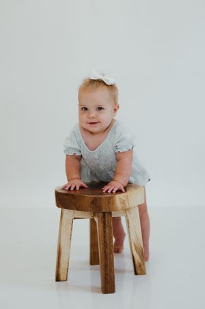 Adorable baby girl in a light blue romper and white bow headband standing at a small wooden stool.