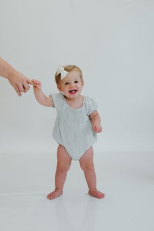 Smiling baby girl in light blue romper with white bow stands barefoot, holding an adult's finger for support.