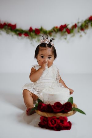 Baby girl in white lace dress enjoys first birthday cake smash with red roses and floral garland backdrop.