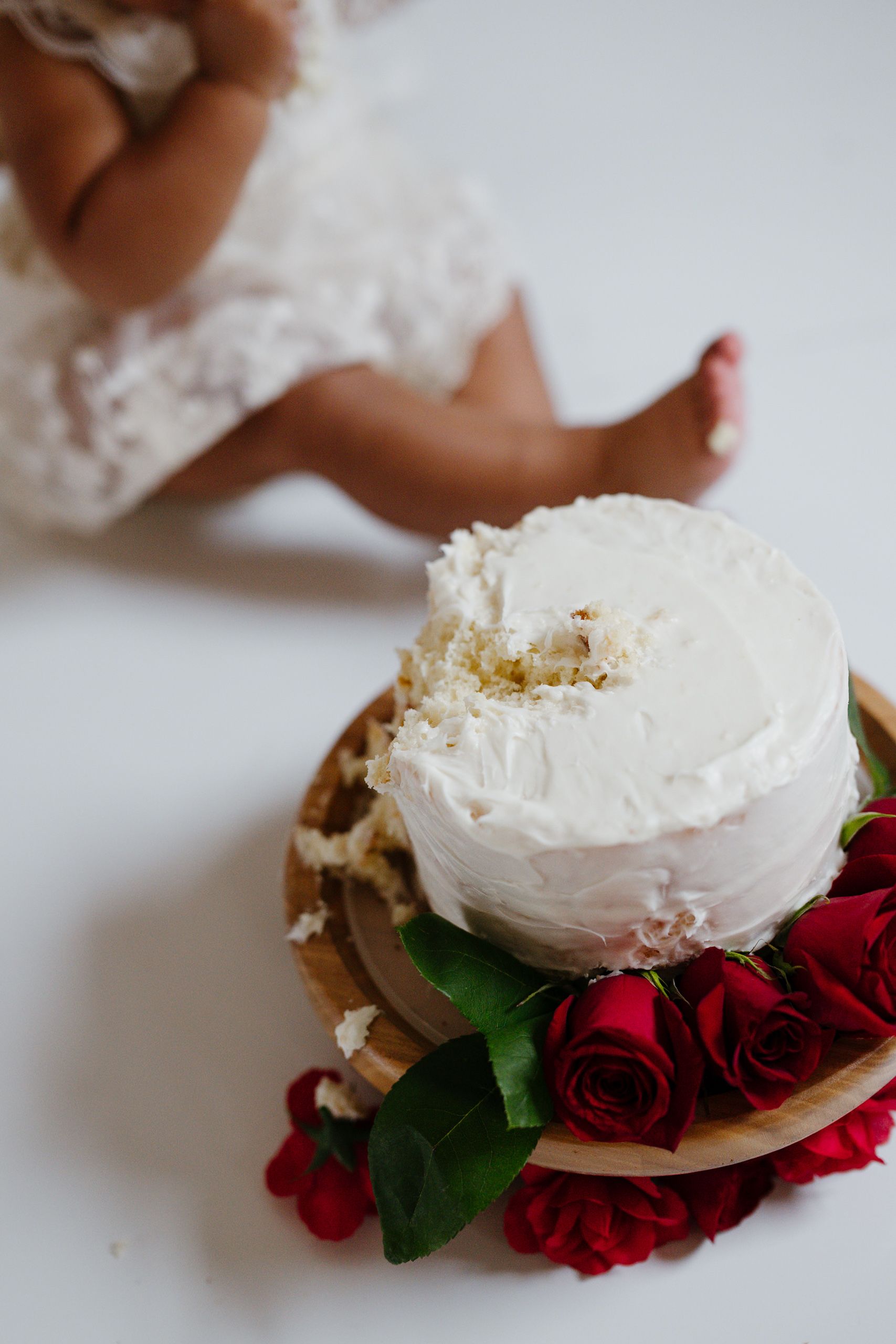 White frosted smash cake decorated with red roses on wooden board, with baby in lace dress in background.