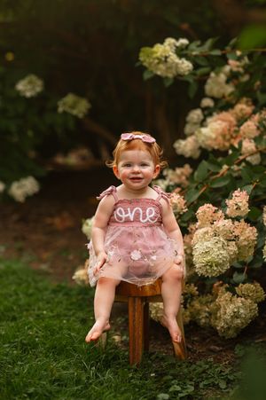 Smiling baby girl in pink tutu romper sitting on wooden stool surrounded by hydrangea blooms for first birthday photos.