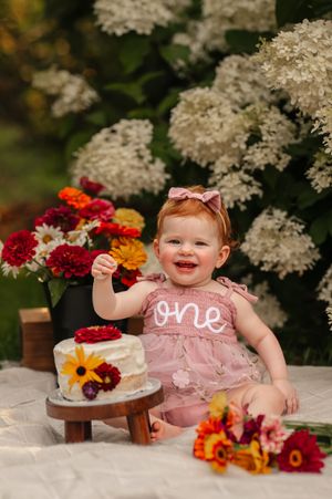 Baby girl in pink "one" dress smiles at first birthday cake smash surrounded by colorful flowers outdoors.