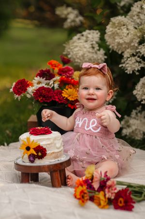A baby girl in a pink 'one' dress celebrates her first birthday with a floral cake outdoors.