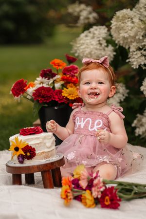 A baby girl in a pink tutu dress celebrates her first birthday with a smash cake outdoors, surrounded by colorful flowers.
