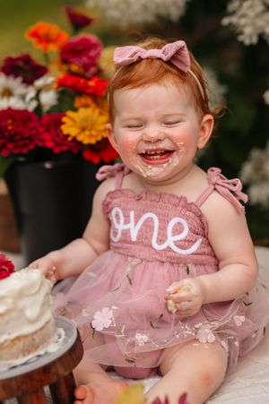 Red-haired baby girl in pink “one” dress laughs with cake on her face during first birthday smash cake photo.