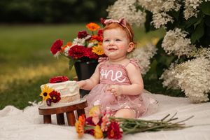Baby girl in pink dress celebrates first birthday with cake and flowers outdoors.