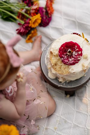 Baby girl in pink tutu reaching for white birthday cake topped with red flower, surrounded by colorful blooms.