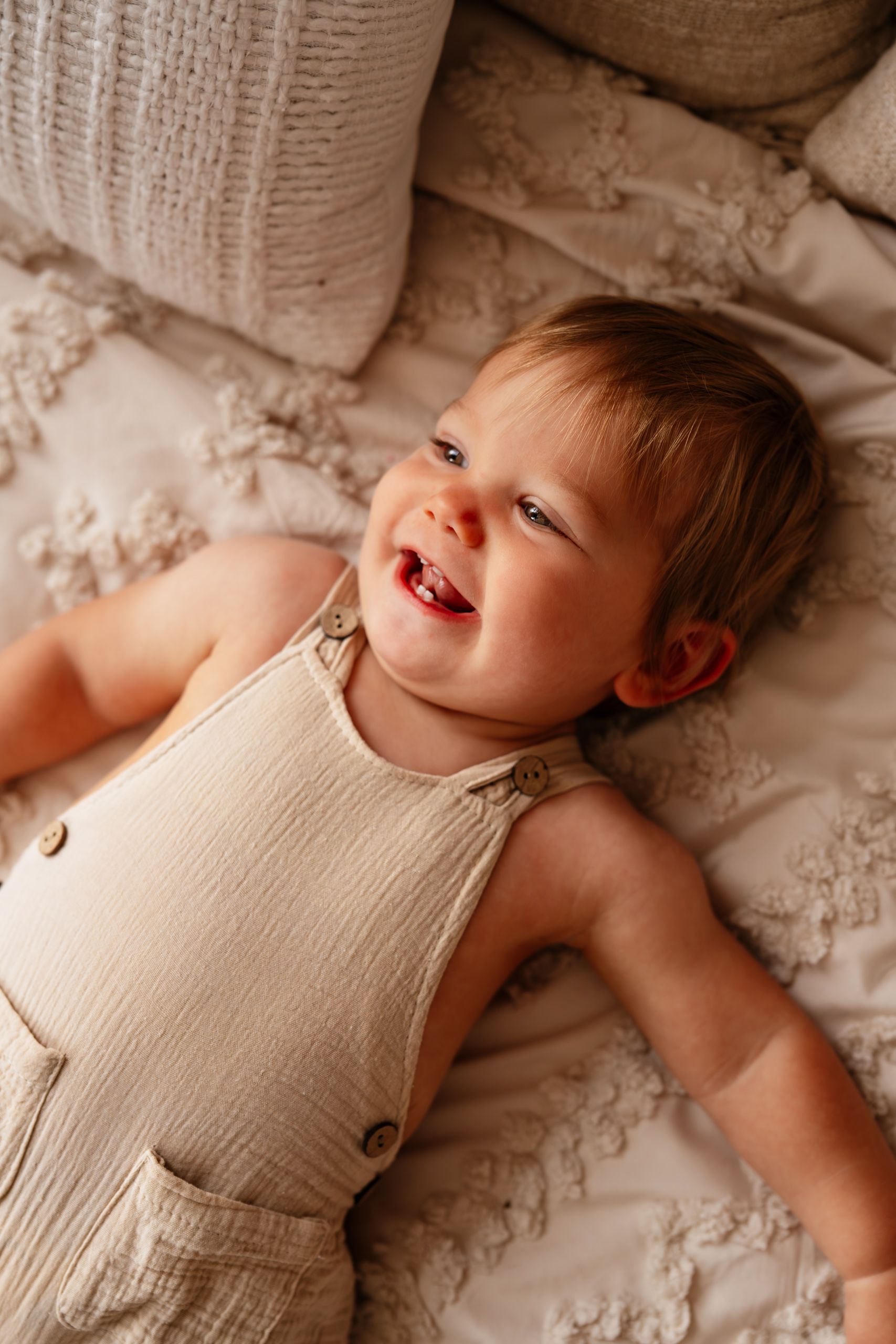 Smiling baby lying on textured beige bedding wearing a cream linen romper.