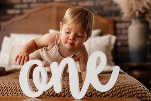 A baby girl celebrates her first birthday sitting on a bed with a white cursive 'one' sign in a cozy room.