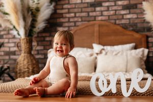 Smiling one-year-old baby sitting on floor beside a wooden 'one' sign celebrating their first birthday.