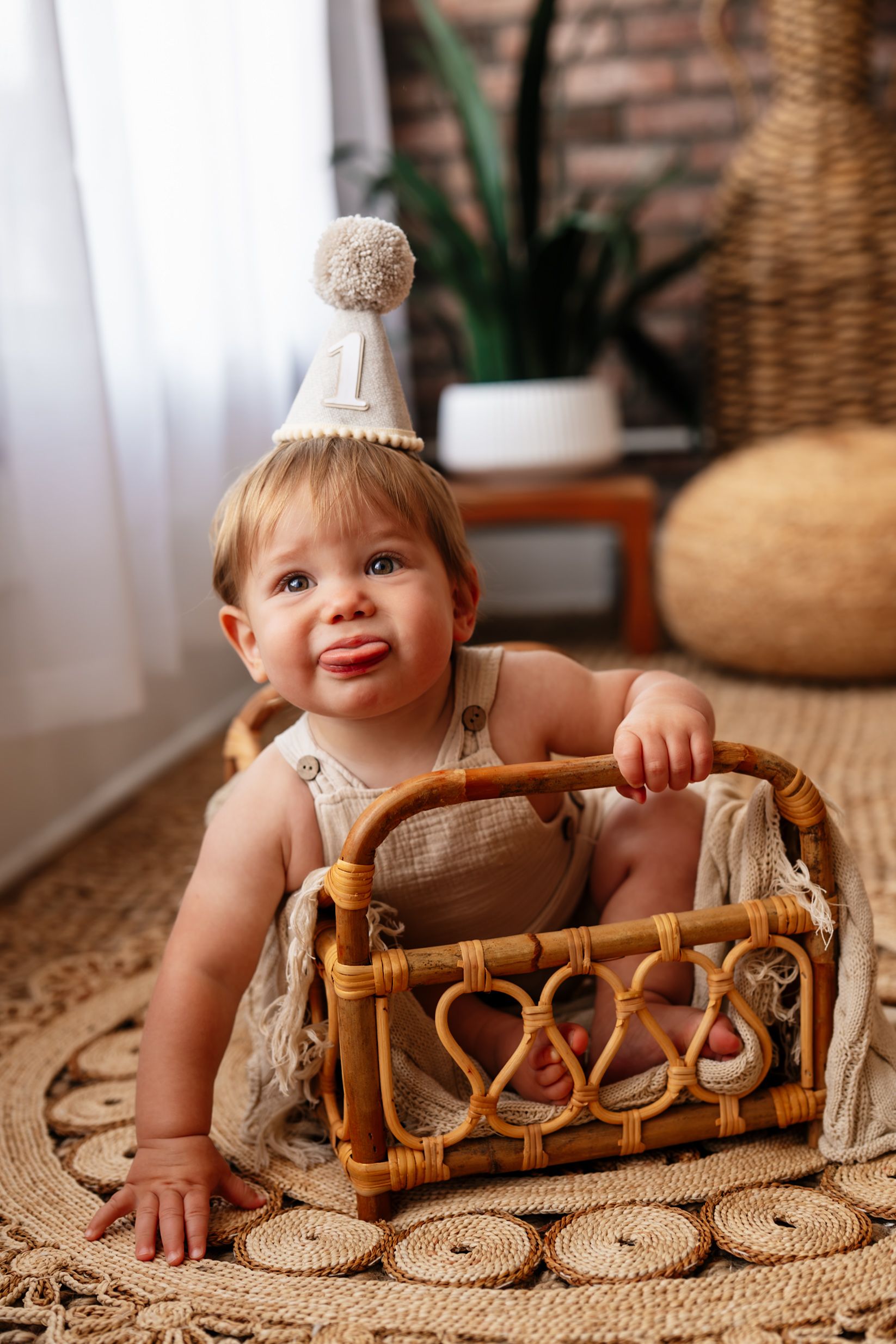 Cute baby wearing a number one party hat sitting inside a wicker basket on a jute rug for first birthday photos.