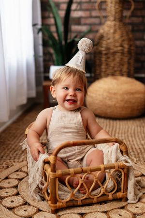 Smiling baby in beige romper and birthday hat sitting in rattan basket on woven rug in boho-style room.