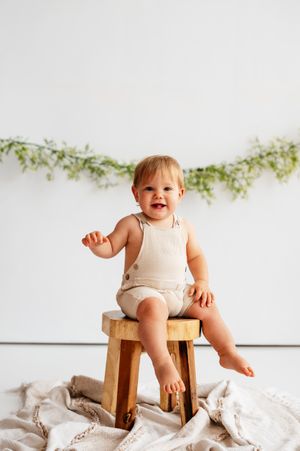 Smiling baby in cream romper sitting on wooden stool with green garland backdrop on white wall.