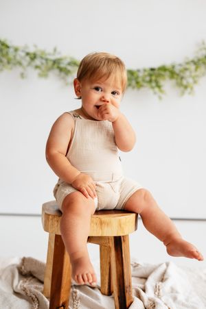 Adorable baby in beige romper sitting on wooden stool with green garland backdrop.