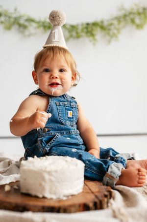Baby in denim overalls and party hat enjoys first birthday cake smash on rustic wood slice.