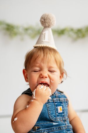 A baby wearing a number 1 birthday hat eating cake with frosting on their face and hand.
