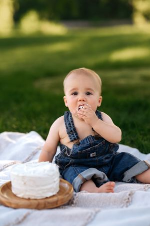Baby in denim overalls eating first birthday cake smash on white blanket in green park.