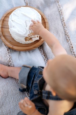 Toddler in jeans reaching into white frosted cake on wooden board.