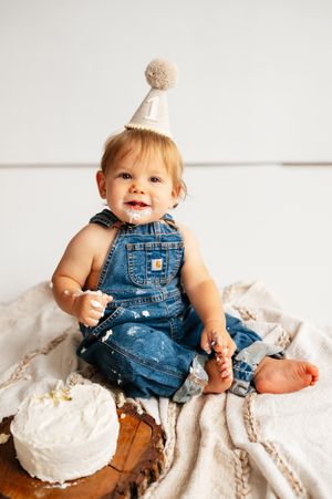 Baby in denim overalls and party hat smiles beside a smash cake during a first birthday photo session.