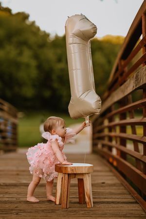 Baby girl in pink dress holds a number 1 balloon on a wooden bridge, celebrating her first birthday outdoors.
