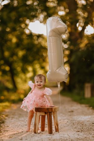 A smiling baby girl in a pink dress sits on a wooden stool outdoors, next to a silver number 1 balloon.