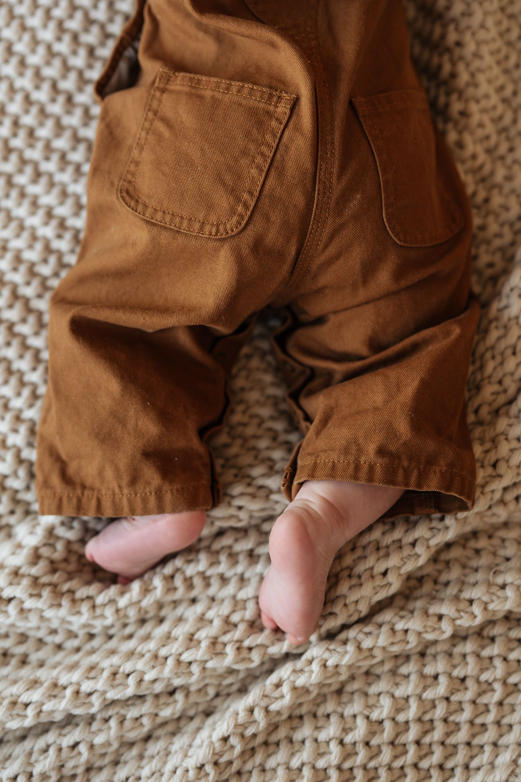 Close-up of baby feet peeking out from brown cargo overalls on a knit blanket.