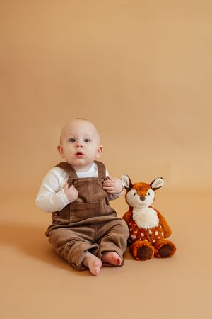 Baby in brown overalls sits with a plush deer toy on a beige background.
