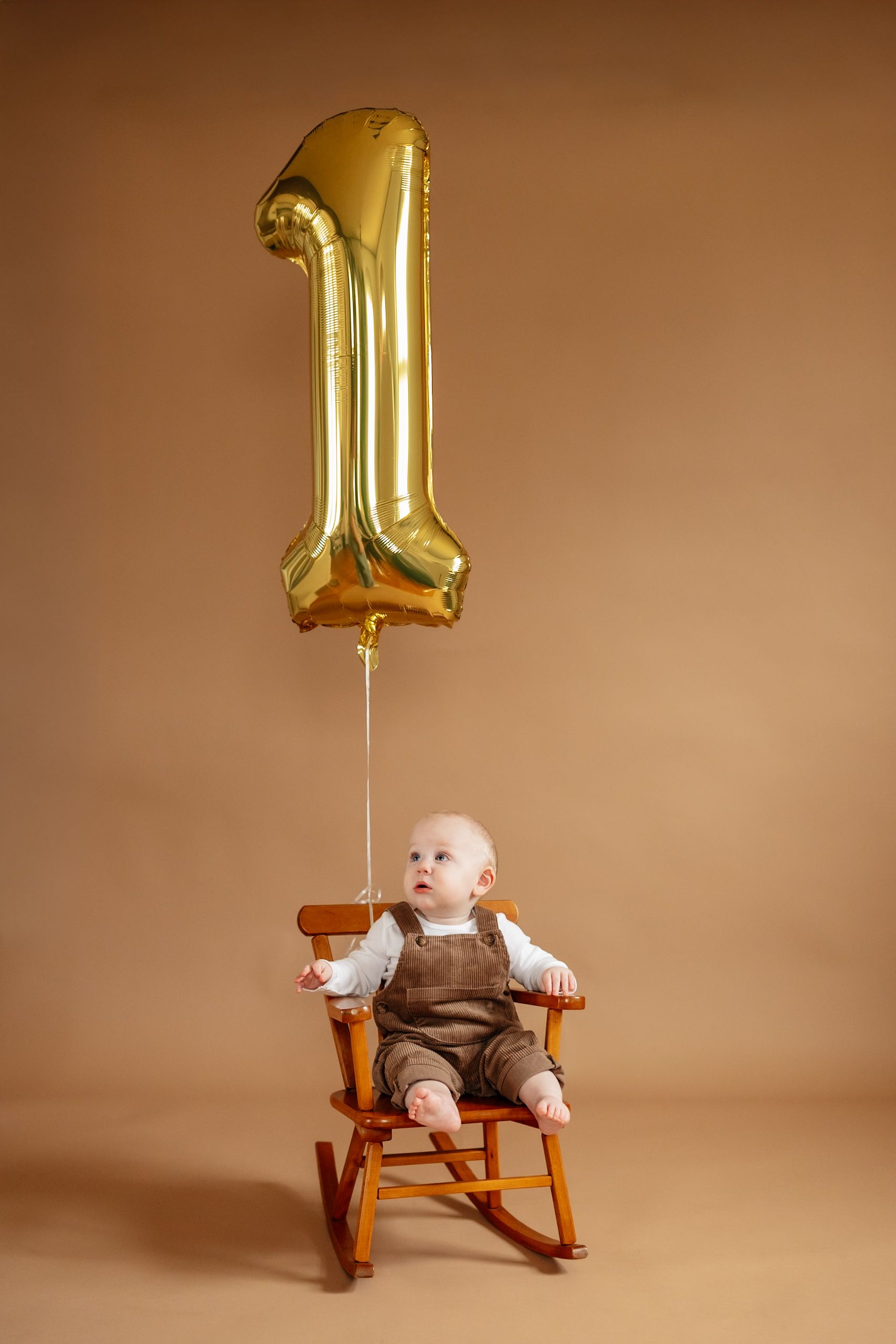 Infant in brown outfit sits in wooden rocking chair with gold number 1 balloon on beige background.