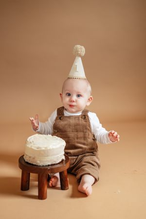 A baby in brown overalls and a number 1 party hat sits beside a white cake on a small wooden stool.