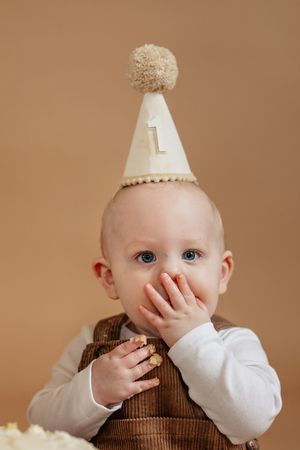 A baby wearing a number one birthday hat eating cake with their hands against a beige background.
