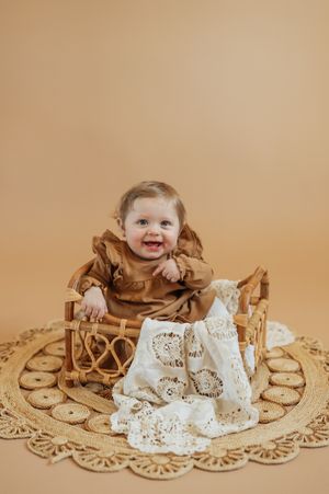 Smiling baby in brown dress sits in a wicker basket on a rattan rug.