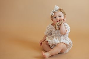 Cute baby girl in checkered romper and bow headband chewing on a wooden teething ring toy.