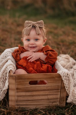 Smiling baby girl in rust orange romper and bow headband sitting in wooden crate outdoors.