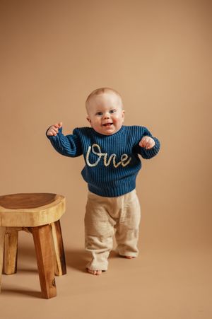 A smiling baby wearing a blue 'One' sweater and beige pants stands next to a wooden stool on a tan background.