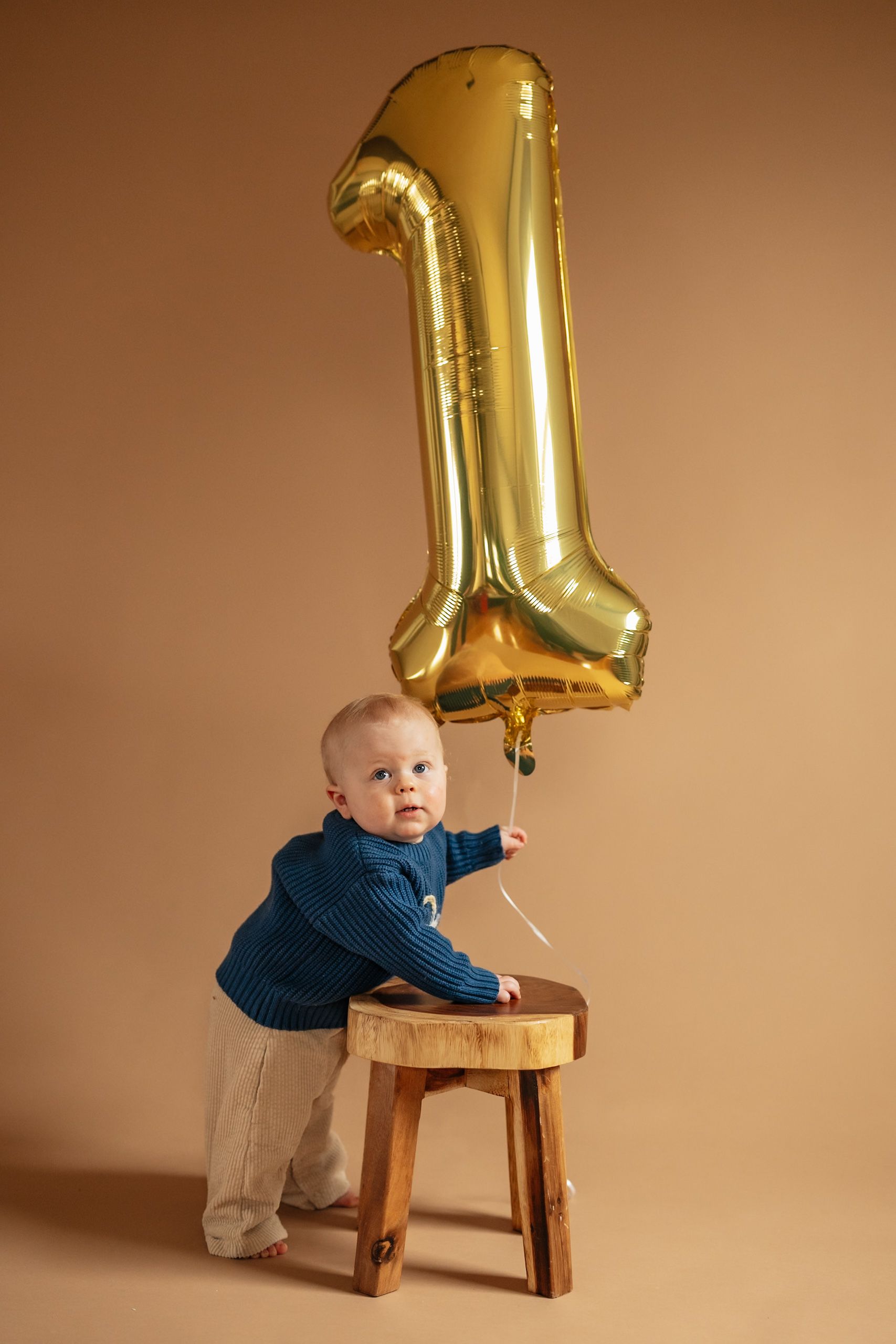 Baby in blue sweater stands at wooden stool holding gold number 1 balloon on beige background.