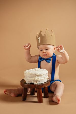 Baby in crown and bow tie celebrates first birthday with white cake on small wooden stool.